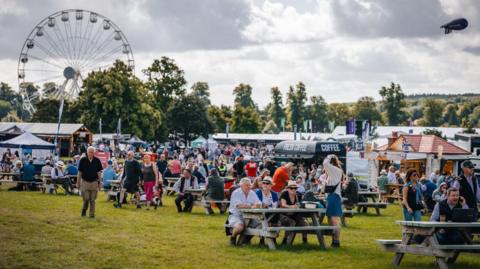 A large gathering of people at an open-air event, with some sitting on wooden picnic benches in the foreground and stalls and a big wheel in the background