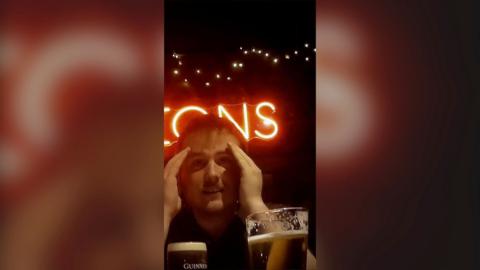 A man with both hands around his face, in a pub, with two pints in front of him and letters in light behind.
