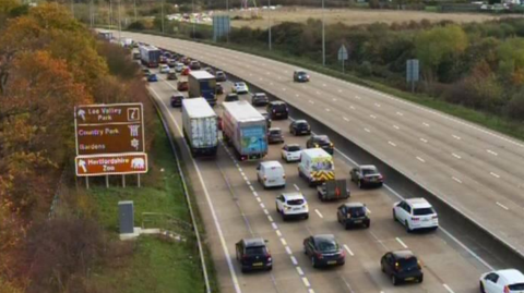A traffic camera image from above part of the M25 in Hertfordshire. Four lanes of the motorway, on the carriageway closest to the camera, are filled with slow moving vehicles. The opposite carriageway is relatively empty apart from one car.