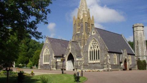 A crematorium. Two small chapels sit either side of the main entrance to the main tower. The buildings are grey stone. to the right is a large grey chimney. To the left is a tree and a patch of grass