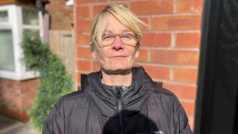 Alison Dhatt has blonde hair and glasses. She is wearing a black coat and standing in front of a house