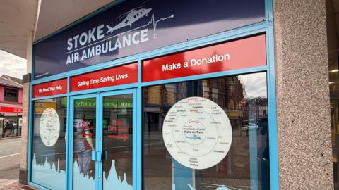 The front of a charity building that has glass doors and windows and red and blue signage. It says "Stoke Air ambulance"