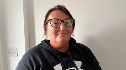 Resident Nicha Rowson, who has brown hair tied back, wears a black hoodie with a white logo. She has dark rimmed glasses. She is standing in front of a white wall and a light switch is over her right shoulder.