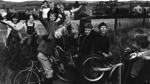 A black-and-white photograph of a group of young boys posing for the camera. A couple are on bikes and the boys above them are crowded together, some with their hands in the air. All of them are smiling and look generally excited.