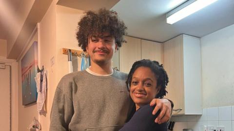 A teenage boy with his arm around his mother, who is hugging him. The teenager is wearing a grey jumper, while his mother is wearing a dark blue top. They are both smiling.