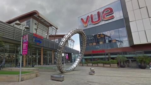 Trinity Square in Gateshead. A large Tesco Extra store stands to the left while a Vue cinema is on the right. A large metal statue, which looks like a metal band leaning over, is in the middle of the square.