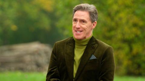 Rob Brydon smiling, wearing green polo neck and green blazer, in front of trees.