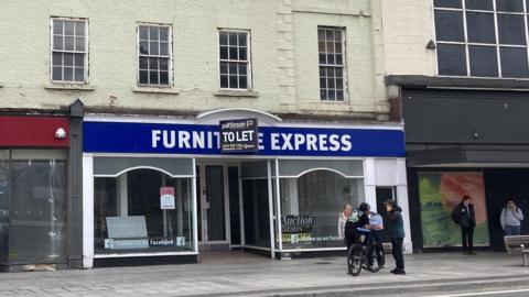 The former Furniture Express store in Stockton. A "To Let" sign is fixed above the doorway. There are large glass windows to either side. A small number of people are chatting on the pavement in front of the building. The upper floors are painted cream.