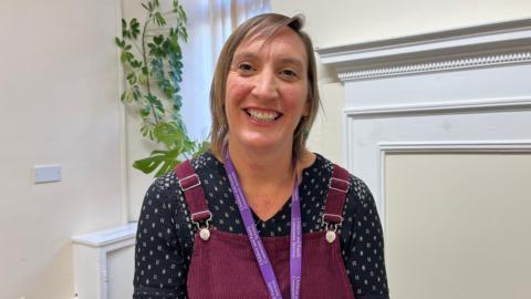 Headshot of Amy Ford in a burgandy pinafore, navy patterned blouse and purple lanyard