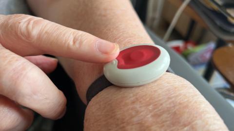 Photo of a red alarm button strapped to a wrist with a black strap with a finger touching it