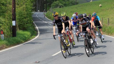 There are several cyclists on the road. To the right and left there is a grassy verge, the cyclists are on the right-hand side of the road.