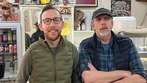 Matt and Barry Howarth standing in the shop. Matt has short dark hair and a beard and black framed glasses and is smiling. His dad has a white goatee and is wearing a baseball hat