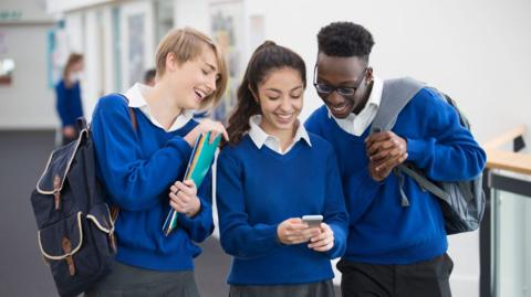 Three pupils wearing blue school jumpers, white shirts and grey skirts/trousers are looking at a phone the girl in the middle is holding while they are walking in the school corridor