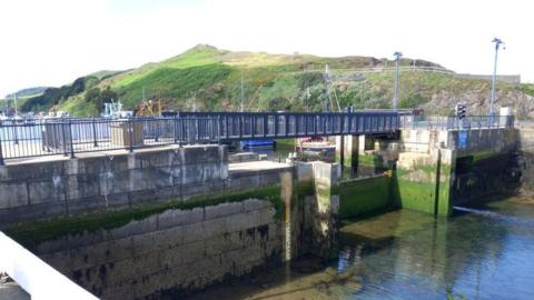 The pedestrian bridge across Peel Harbour at low tide. It has black railings on either side and sits above the harbour walls which have green algae growing on them. Peel Hill sits in the background.
