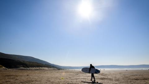 A surfer holding a longboard under his arm walks across a large empty beach. The sky is clear and blue and the sun is shining. The sea is glittering in the distance.