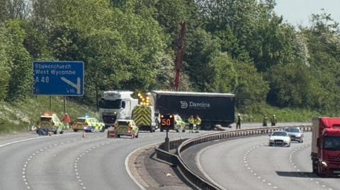 A crane is being used to lift the carriage of a lorry. It is on a motorway surrounded by police cars near a road sign for Stokenchuch and West Wycombe