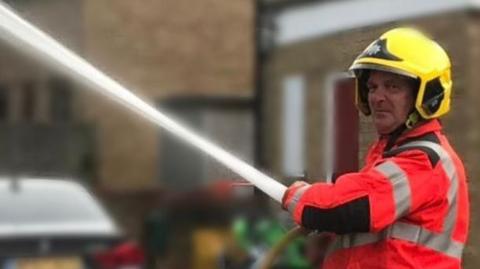 Mervyn Housden wearing a yellow firefighters' helmet, a high-vis orange jacket with grey stripes and operating a water hose