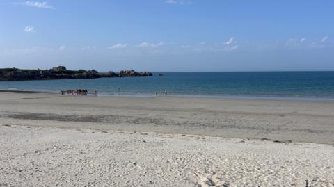 The image is of the beach where you can see the blue sky with clouds, the sea water, some land and rocks on the left, and sand. 