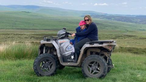 Katherine Singer is sitting on a quadbike with her daughter Sophie. Behind them is a large area of Northumberland uplands with no houses visible anywhere 