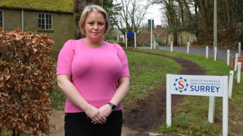 A woman wearing a pink top, stood in front of an area of grass. She is looking directly at the camera. 