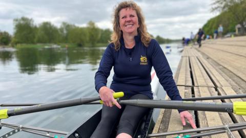 Kate Lindgren wearing a blue fleece sitting in a rowing boat holding her oars. Her left hand is leaning on wooden decking, and in the left of the photo and in the background is the river and some trees on the bank.