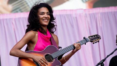 Olivia Dean wears a bright pink dress and plays the guitar, which is on a strap hanging around her. She has long curly black hair and wears gold rings and bracelets