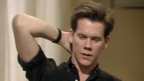 Kevin Bacon holds his hand behind his head.