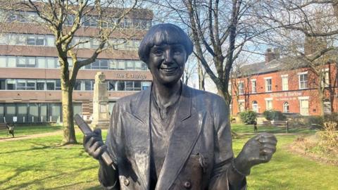 A bronze statue of Victoria Wood, holding her arms apart and smiling, stands in a garden by a public library on a sunny day.