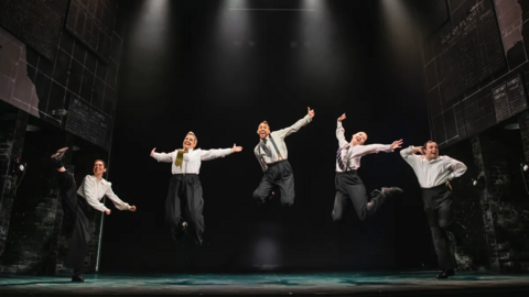 Five actors dressed in white shirts and grey trousers stand on stage while jumping in the air.