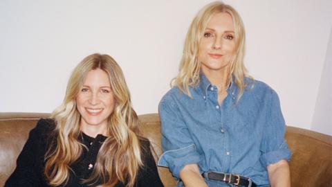 Two women with blonde hair smiling, one wearing a black jacket and the other a blue button up shirt.