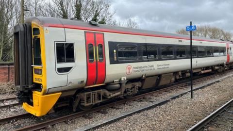 The front of a Transport for Wales train, which is grey and red, on a railway line. The sky is grey in the background. 