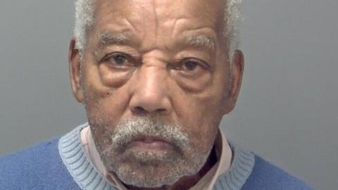 Headley's custody picture. He has white hair, eyebrows and short facial hair, and wears a white collared shirt and a blue jumper. He is looking straight ahead with a serious expression. 