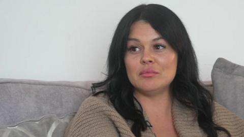 Jody, who is wearing a beige cardigan, is sitting on a grey couch during an interview. She is staring off camera. 