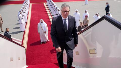 Keir Starmer boarding a plane at the airport in Doha, Qatar. There is a red carpet, lined with men, leading up to the plane