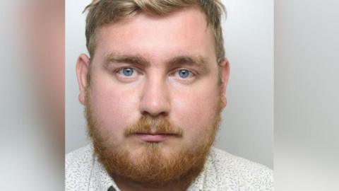 Headshot of man with ginger beard and moustache and light brown hair with blue eyes wearing a cream and flowery shirt