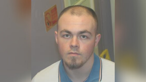 A man with thinning black hair and a beard is looking at the camera. He is wearing a white top which has a blue collar.