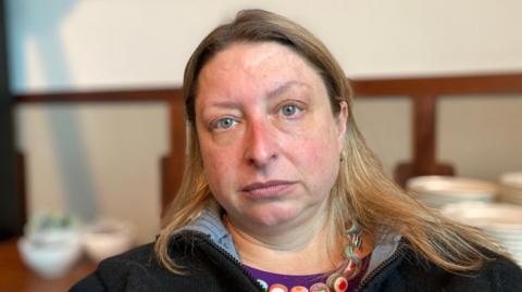 A woman with long brown/blonde hair looks straight ahead, unsmiling. She wears a dark zipped up top and a decorative necklace.
