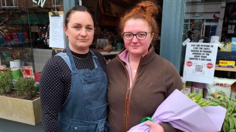 Ruth Phillips (left) and Charlotte Kneafsey. Ruth is on the left has her hair tied back and is wearing denikm dungarees with a black shirt with white spots. Charlotte has red hair tied up, black glasses and is wearing a brown flees. She is holding a bunch of flowers wrapped up. They are standing in front of a shop.