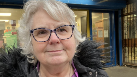 A woman stands in front of the glass doorway to a large public library. She has white hair and wears glasses and a parka-style coat.