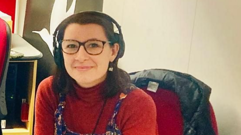 A picture of Jayne Jackson, who is wearing headphones in the radio studio, wearing a red jumper and purple dungarees, sitting on a red chair with a black coat 