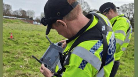 A police officer wearing a yellow hi-vis jacket operating a drone in a field. He is looking at the control screen display.