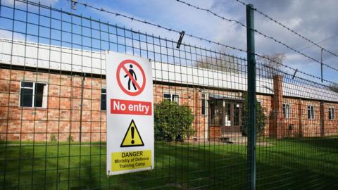 A general view of Crowborough Training Camp, East Sussex. There is a wired fence and a sign, with an area of grass behind it. 