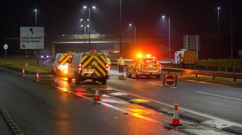 The road closure on the A66 following the crash. Two police officers stand on the road next to a police car. Two National Highways vans are parked next to it.  A police van can be seen on the opposite carriageway.