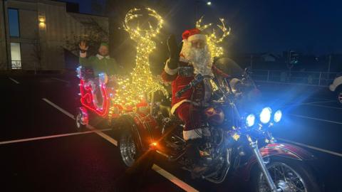 A man dressed in a Santa suit is sat on a red glittery motorcycle with three wheels. Behind him is a sleigh that has been illuminated in red lights with a man in a green elf costume sat inside it. Between the two men are two illuminated golden reindeer. 