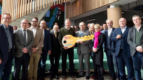 A group of representatives from a council and businesses stand together inside a new innovation centre. Two people in the middle hold a large yellow-coloured cardboard key.