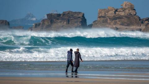 Couple walk along a beach with crashing waves in the background