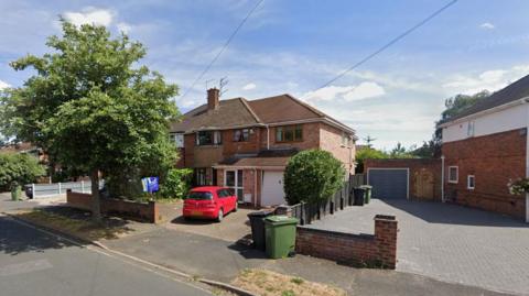 A house with a red car in the front of the drive. There is a large tree on the pavement outside of the house.