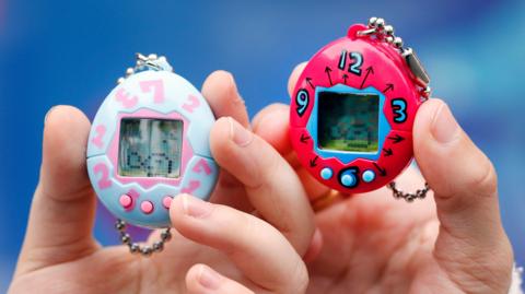 Two children's hands holding tamagotchis. They are egg shaped, palm sized electronic toys. There is a screen with three buttons beneath it. On the left, the tamagotchi is light blue with light pink buttons at the bottom and light pink numbers around the screen. On the left is a bright pink tamagotchi with bright blue details.