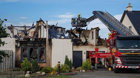 The burned out remains of a suburban house, with a fire engine stationed on the left of the property. Firefighters are examining the scene, one of whom is on to of a height appliance.