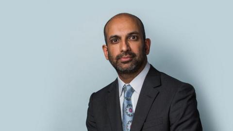 Rajiv Menon KC, who has a beard and brown eyes, standing in a grey suit in front of a blue wall.